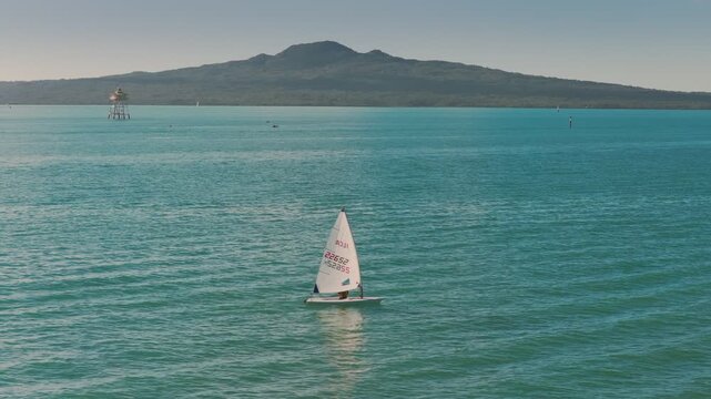 AUCKLAND, NEW ZEALAND - 16 June 2026:  A lone sailor navigates the calm waters of the Hauraki Gulf near Auckland, New Zealand, with Rangitoto Island in the background.