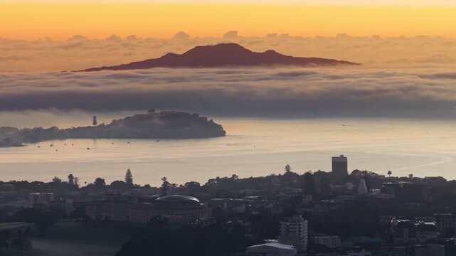 Auckland, New Zealand - Sunrise over the Hauraki Gulf, with Rangitoto Island shrouded in mist and low clouds, creating a serene, ethereal atmosphere.