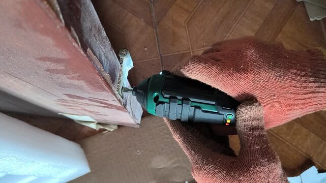 With nobody else around, gloved hands carefully use a compact cordless electric screwdriver to repair a wooden door hinge during a home renovation, evoking feelings of focus.