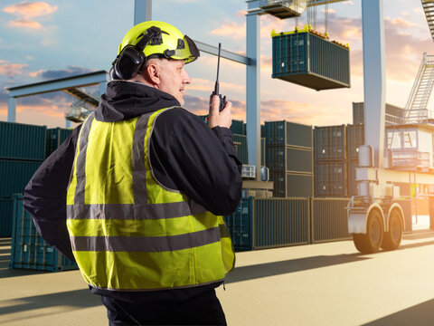 Logistics terminal, Container port. Dock worker uses radio to coordinate crane loading and cargo container handling at shipping yard