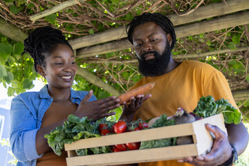 Obraz premium African couple standing under pergola holding wooden crate with sweet potato, tomatoes, kale, beets