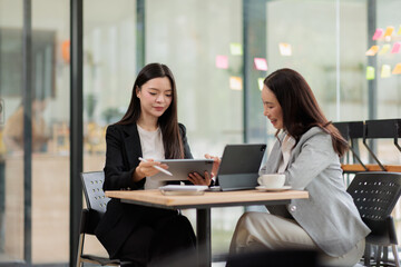 Fototapeta premium Asian businesswomen collaborating on a digital tablet, discussing project ideas and strategy in a modern office meeting, demonstrating teamwork and digital innovation