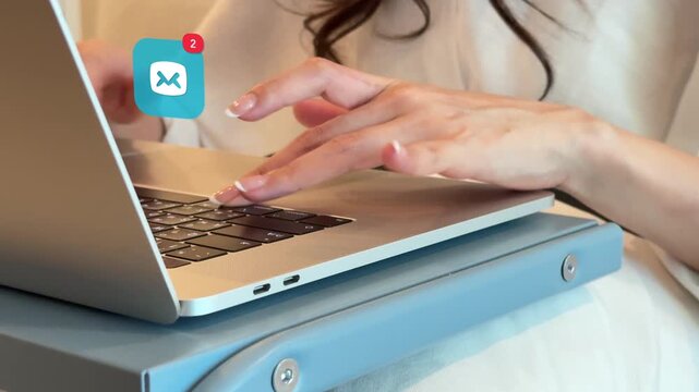 Close up of a young East Asian woman typing on her laptop keyboard, with a digital mail envelope message alert bubble popping up on screen.