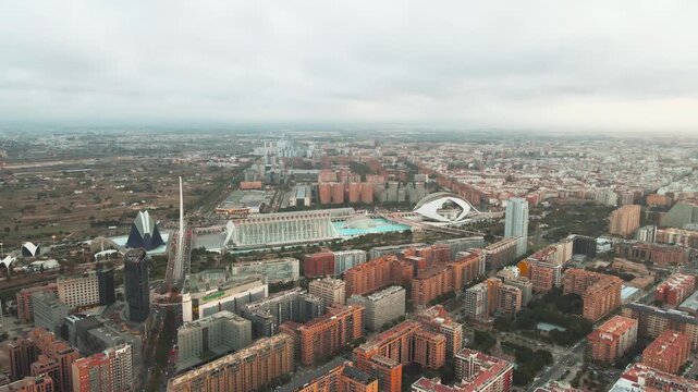 Aerial view of a bustling cityscape. Modern skyscrapers and red-brick buildings coexist. Prominent curved roof structure dominates the skyline. Overcast sky casts soft light over the area