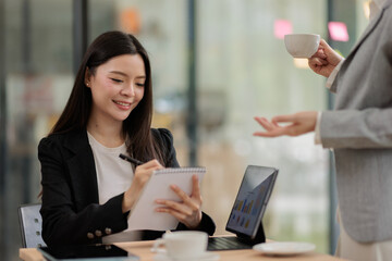 Obraz premium Asian businesswoman smiling and writing notes in a spiral notepad during a business meeting, actively listening to a colleague and collaborating on data analysis