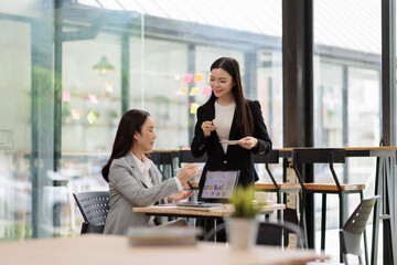 Obraz premium Two young asian businesswomen collaborate in a modern office, reviewing financial charts on a laptop while sharing ideas over coffee, focused on strategy and growth