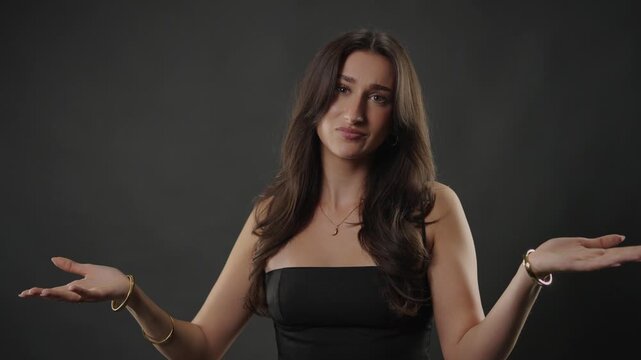 Uncertainty and confusion concept, a young woman shrugging with open hands and a puzzled expression on a dark studio background. This footage doubt, hesitation, and the feeling of not knowing, commerc