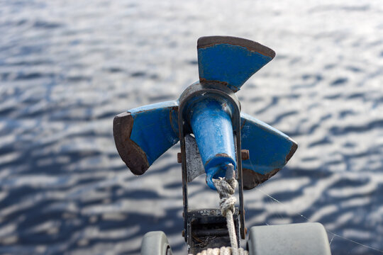 boat anchor mounted on a deck bow roller assembly. detailed marine safety hardware texture showing weathered iron anchor flukes, a steel shackle hook, and a rippling water background under daylight.