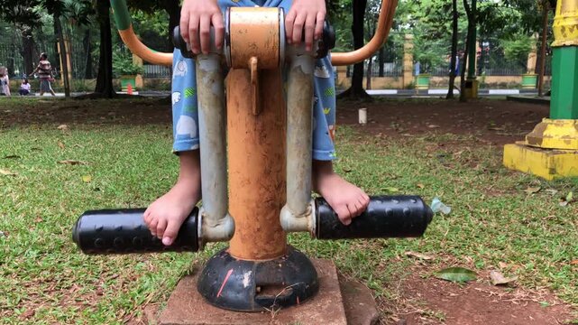 Barefoot child in blue pajamas playing on outdoor gym equipment at a public park surrounded by green trees