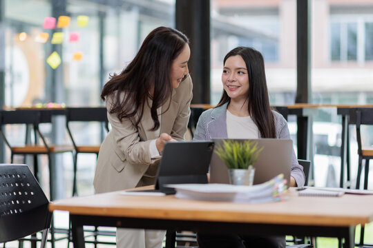 Two smiling asian businesswomen engaging in a joint project on a laptop, sharing knowledge and working together at a desk in a contemporary office space