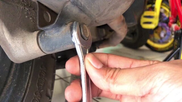 Close-up of a mechanic's hand using a metal wrench to tighten a bolt on a dusty motorcycle exhaust pipe during repair work.