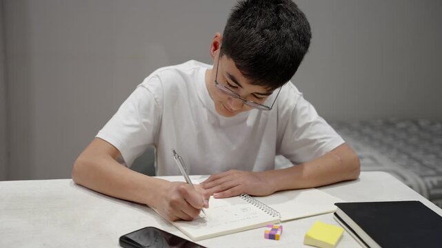 Front view of teenage student in glasses writing in notebook at home desk with stationery. Focused learning, homework, education, study routine and school preparation concept.