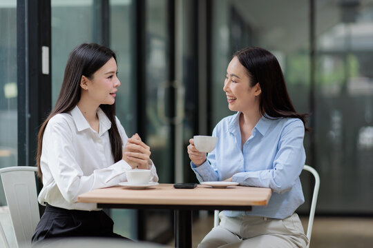 Two asian businesswomen discussing work outdoors, enjoying coffee, smiling, collaborating, engaging in networking, and having a productive meeting at a cafe table