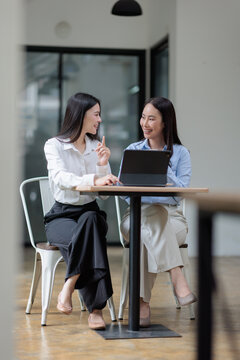 Two young asian business women collaborating on a project and communicating ideas, discussing work using a tablet computer in a modern coworking office space