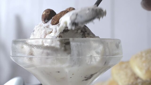 Vanilla ice cream with chocolate in a glass cup, with almonds on top, illuminated by window light on a summer afternoon and a rustic white wooden wall in the background. grabbing with a spoon