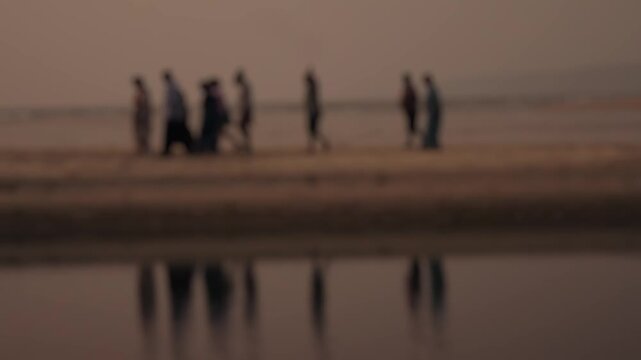 Unfocused, dreamy shot of a group of people walking along the shoreline at dusk, with their reflections cast in the wet sand, creating a serene and melancholic atmosphere