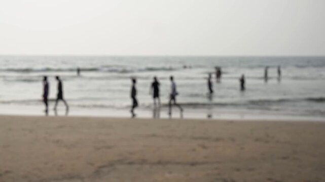 Defocused silhouettes of anonymous people enjoying a sunny day at the seashore, walking on the wet sand and bathing in the calm ocean waves, creating a peaceful and relaxing summer vacation scene