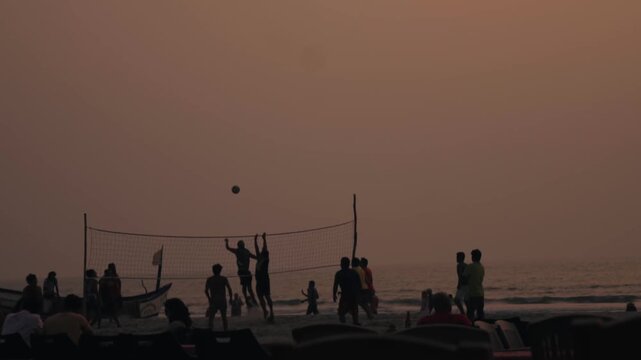 Silhouettes of a group of friends playing volleyball on a sandy beach against the backdrop of the ocean during a hazy and warm sunset, enjoying an active and recreational vacation