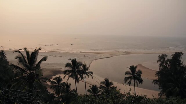 Beautiful panoramic aerial view of a serene tropical beach at dusk, with gentle waves lapping the shore and silhouettes of palm trees against the hazy, warm sky in the foreground