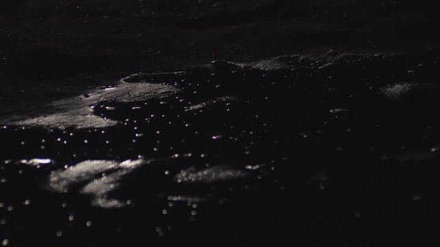 Glistening moonlight reflects and dances on the surface of a dark, mysterious liquid, creating an abstract and cinematic bokeh effect with gentle ripples and shimmering highlights in the night