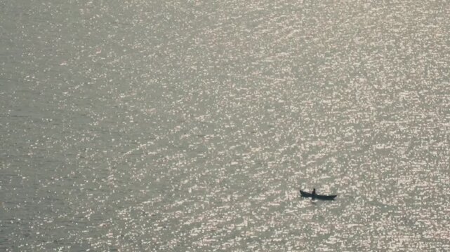 Aerial view of a solitary fisherman paddling a traditional wooden canoe across a vast expanse of shimmering water, with the sun's reflection creating a beautiful sparkling effect on the surface