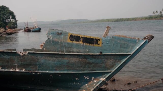 Weathered and broken wooden fishing boat resting abandoned on a muddy riverbank, with other traditional vessels anchored in the background near a lush, tree-covered shoreline