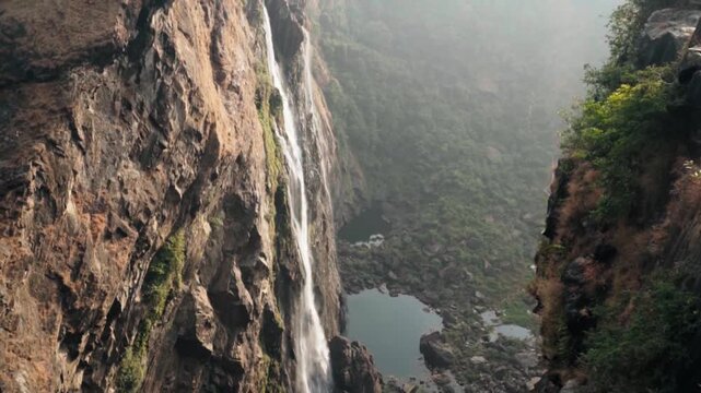 Scenic perspective of a tall waterfall cascading down a sheer rock cliff into a deep river gorge, with the camera tilting upwards to reveal the vast, hazy jungle landscape of Karnataka, India