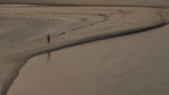 Lone woman strolling on a narrow sandbar, enjoying the serene and tranquil atmosphere of the deserted beach with calm water reflecting the muted tones of the sky in a peaceful landscape