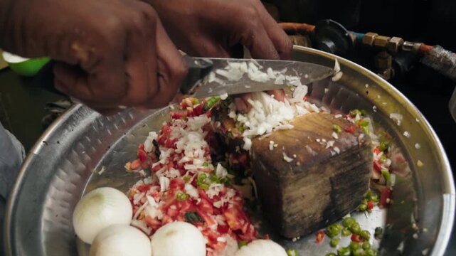 Professional chef's hands chopping onion, tomato and chili peppers on a wooden board over a metal bowl, preparing fresh ingredients for traditional Indian street food