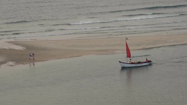 Aerial view of a small tourist boat with a vibrant red sail navigating a calm channel while two people enjoy a leisurely walk along the sandy shore near the ocean waves in Goa, India
