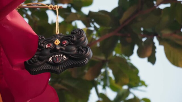 Close-up of a fearsome black demon mask with red eyes and sharp fangs, hanging from a string and gently swaying outdoors with a red flag and green tree leaves in the background