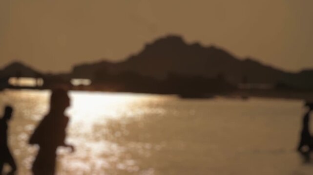 Defocused silhouettes of people walking and enjoying the shallow water of the sea at sunset, with the sun reflecting on the calm surface and mountains in the background creating a serene scene