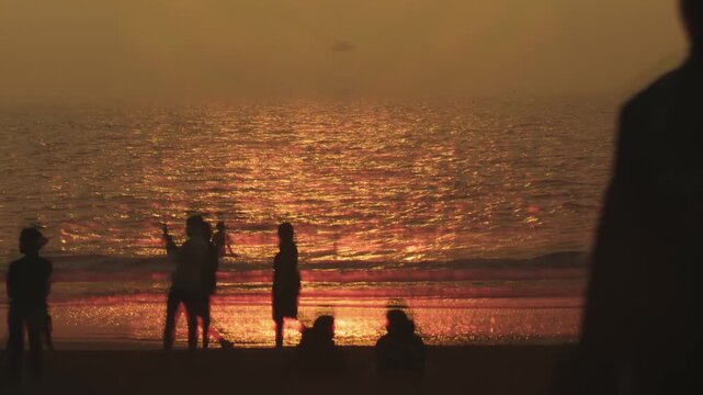 Anonymous silhouettes of people enjoying a beautiful golden sunset on a sandy beach, with the orange sun reflecting on the calm ocean waves as they stand, walk, and sit along the shoreline