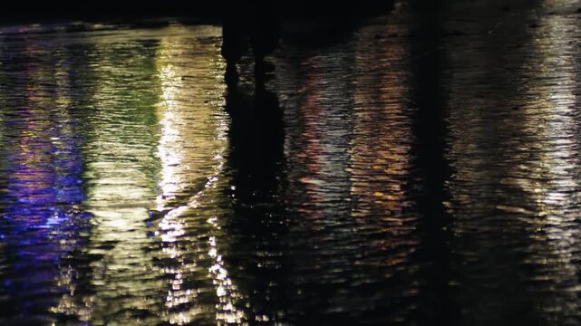 Reflective view of a person's legs walking on a wet urban pavement at night, with colorful city lights shimmering and creating a vibrant, moody atmosphere on the watery surface