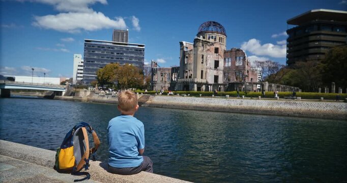 Child Sits by River Facing Hiroshima Atomic Bomb Dome In Japan - Wide, Slow Motion