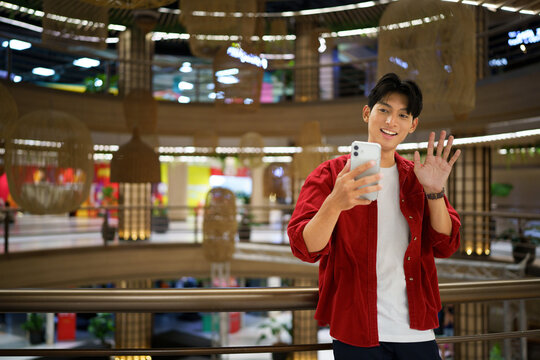 Young Asian Man Waving During Video Call on Smartphone in Modern Shopping Mall