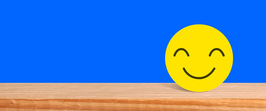 Yellow smiling face icon on wooden table against blue background perfect for positive customer feedback and satisfaction concepts