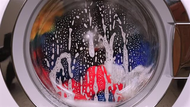 Washing machine glass door as the drum spins rapidly, creating dynamic foam patterns and water motion. Concept of laundry process, cleaning cycle, household routine, and detergent action