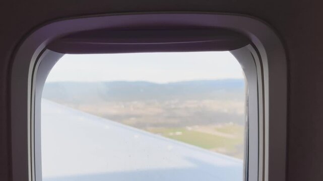 Airplane wing seen through a passenger window during descent over hazy hills and countryside, showing soft in-flight travel scenery.