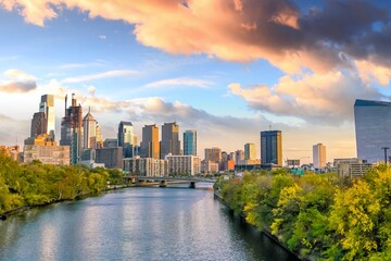 Fototapeta premium Scenic view of the Philadelphia skyline along the Schuylkill River at sunset, featuring modern skyscrapers, vibrant clouds, lush riverside foliage, and reflections in calm water.