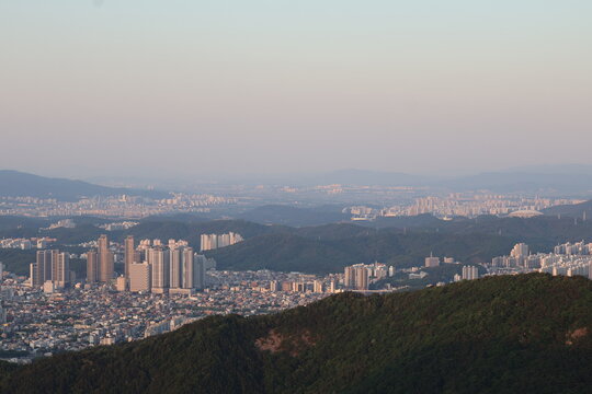 A sunset cityscape view of Daegu, South Korea, seen from Apsan Mountain, with green hills in the foreground and distant urban buildings under a soft evening sky.