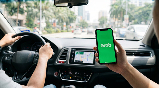 Person holding a smartphone with the green Grab ride-hailing app logo on the screen inside a car cabin with a driver, steering wheel, and GPS navigation system on the dashboard background.