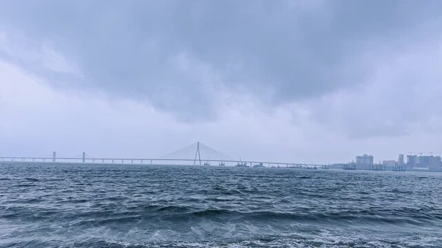 View of Bandra Worli Sea Link from Dadar Chowpatty in Mumbai during a rainy monsoon day with dark storm clouds over the Arabian Sea and city skyline in the distance.