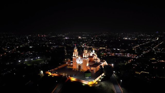 Cinematic nighttime drone shot captures the yellow Cholula church glowing like a beacon atop the shadowy Tlachihualtepetl pyramid below in cholula mexico.