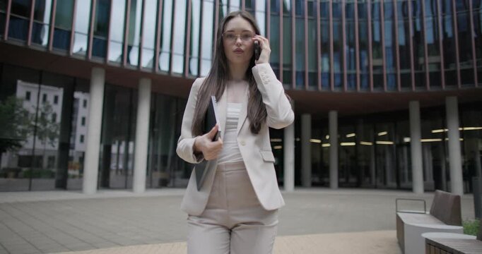 A young Caucasian businesswoman or student or intern in a business suit and glasses stands in an urban setting among trees and modern buildings, holding a laptop and talking on the phone