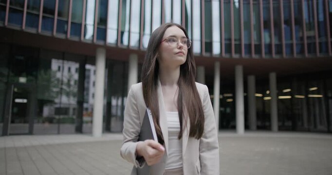 A young Caucasian businesswoman, student, or intern in a business suit and glasses walks down a street among modern buildings, holding a laptop and looking at the camera.