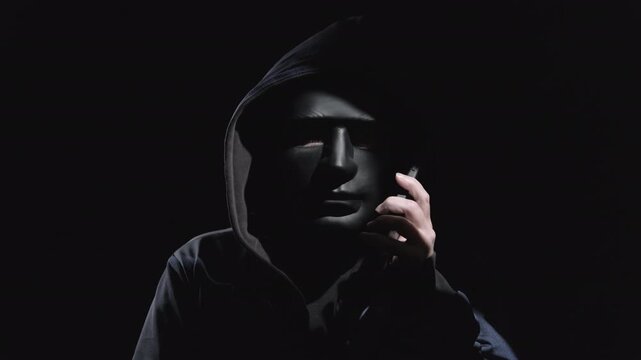 Man scammer wearing black mask on face using smartphone to commit fraud is talking on phone on dark background