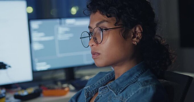 Close-up young Black female programmer coding AI software on multiple screens in night office. Network security, cybersecurity, machine learning, working late, overtime, robotics automation concept.