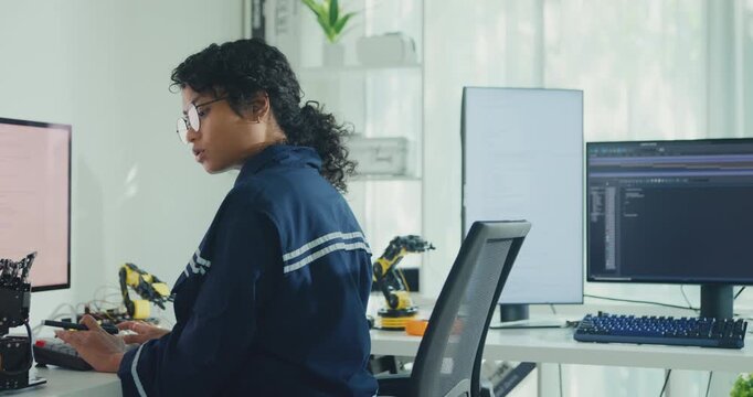 Young African American female robotics engineer coding AI software for robotic arm in laboratory. Embedded system, firmware testing, machine learning, industrial automation, future technology concept.