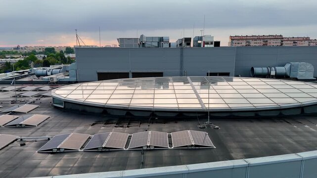 Wide shot of a modern flat commercial building rooftop featuring a large circular glass skylight, solar panels, extensive ventilation system, industrial HVAC units, and residential blocks under a clou